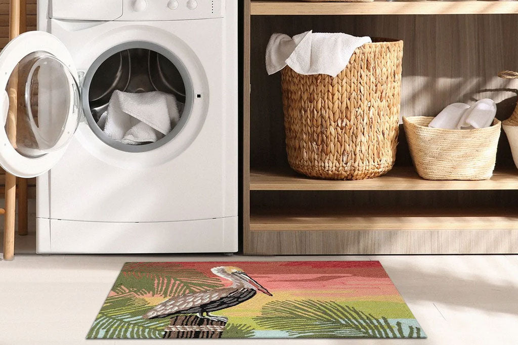 Indoor/Outdoor rug with pelican perched on a wooden post surrounded by tropical green leaves on a bright orange and pink sunset background- shown infront of washing machine in laundry room