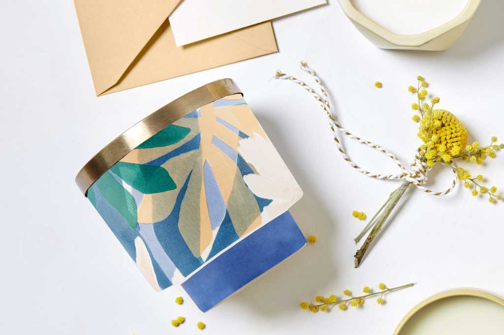 porcelain candleholder with metal lid and candle interior. printed with modern palm-style graphic and lid is gold. Shown on desk with other candles and stationery