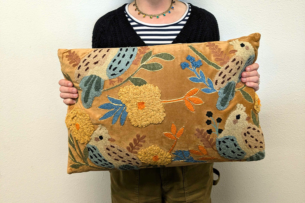 Person holding a decorative pillow with floral and animal patterns against a plain background