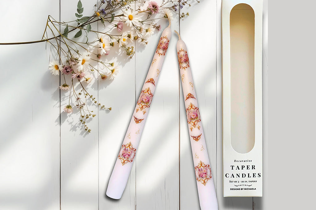 Decorative taper candles with floral design on a white wooden surface with flowers.