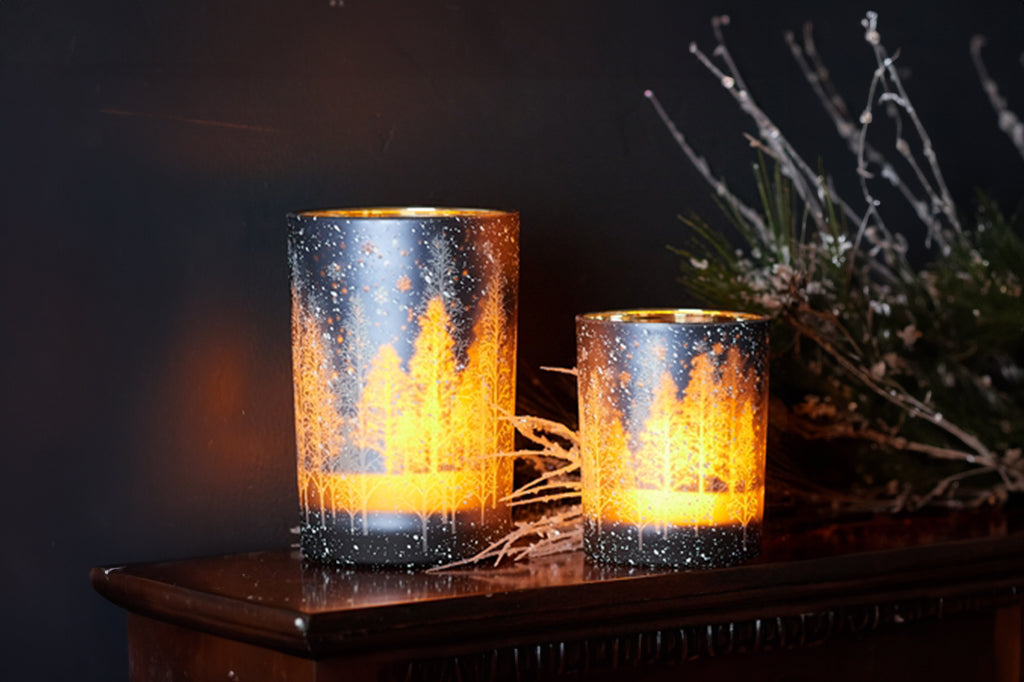 2 candleholders with printed, multi layer tree design on glass shown with lit candles glowing from within; displayed on holiday fireplace mantle