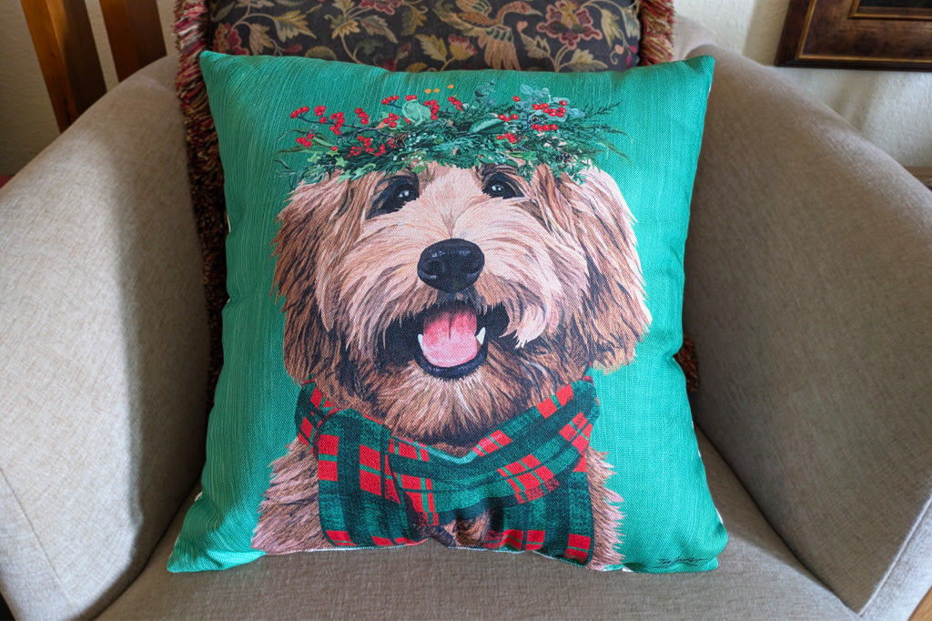 A square decorative throw pillow featuring a printed illustration of a fluffy golden dog with a green wreath and red plaid scarf, set against a turquoise background and displayed on a beige armchair.
