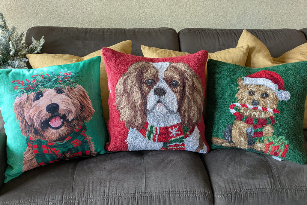 Three decorative throw pillows featuring different dog portraits in holiday colors, arranged on a dark gray couch with yellow pillows behind them.