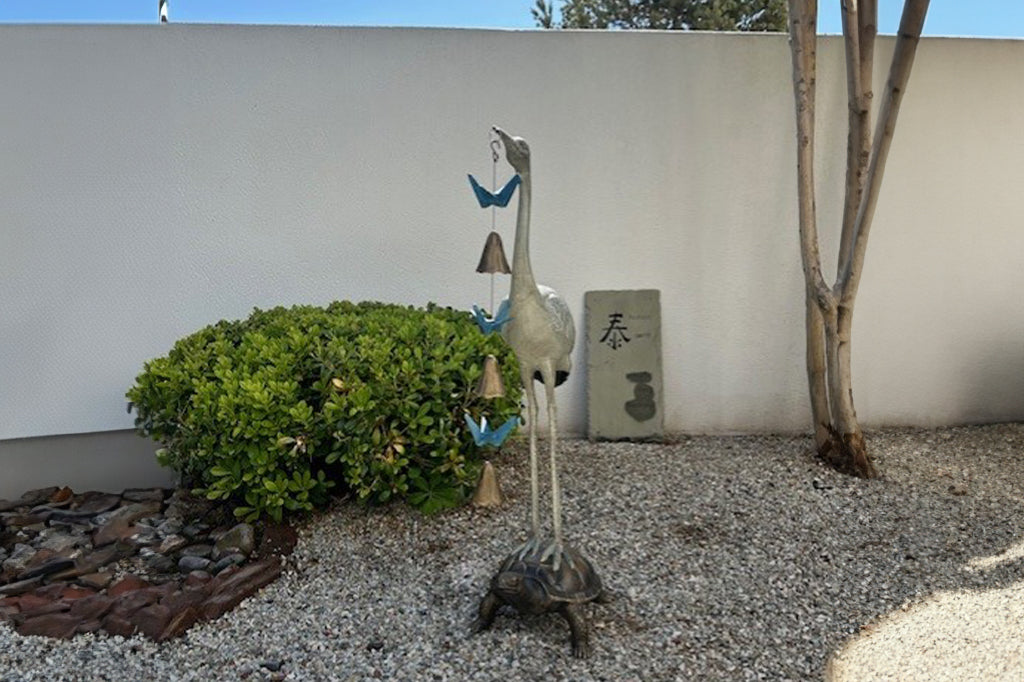 Crane and turtle metal sculpture with bell and blue origami cranes set in rock garden 