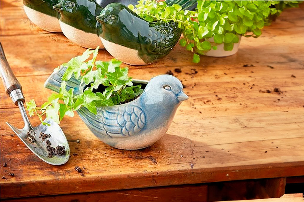 Bluebird Shaped ceramic planter in glossy crackle glaze of cream and watery blue.  Shown on potting table next to a spade, spilled soil, and assorted plants.