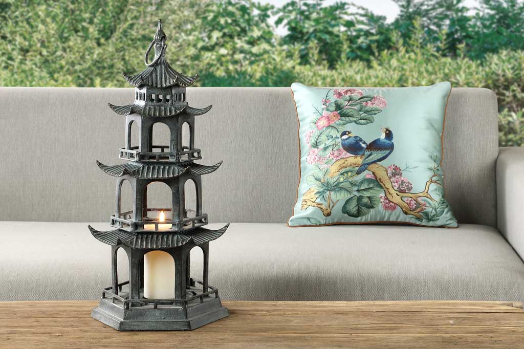 sculptural cast metal lantern shaped like Chinese pagoda holding pillar candle shown outside by sofa and trees
