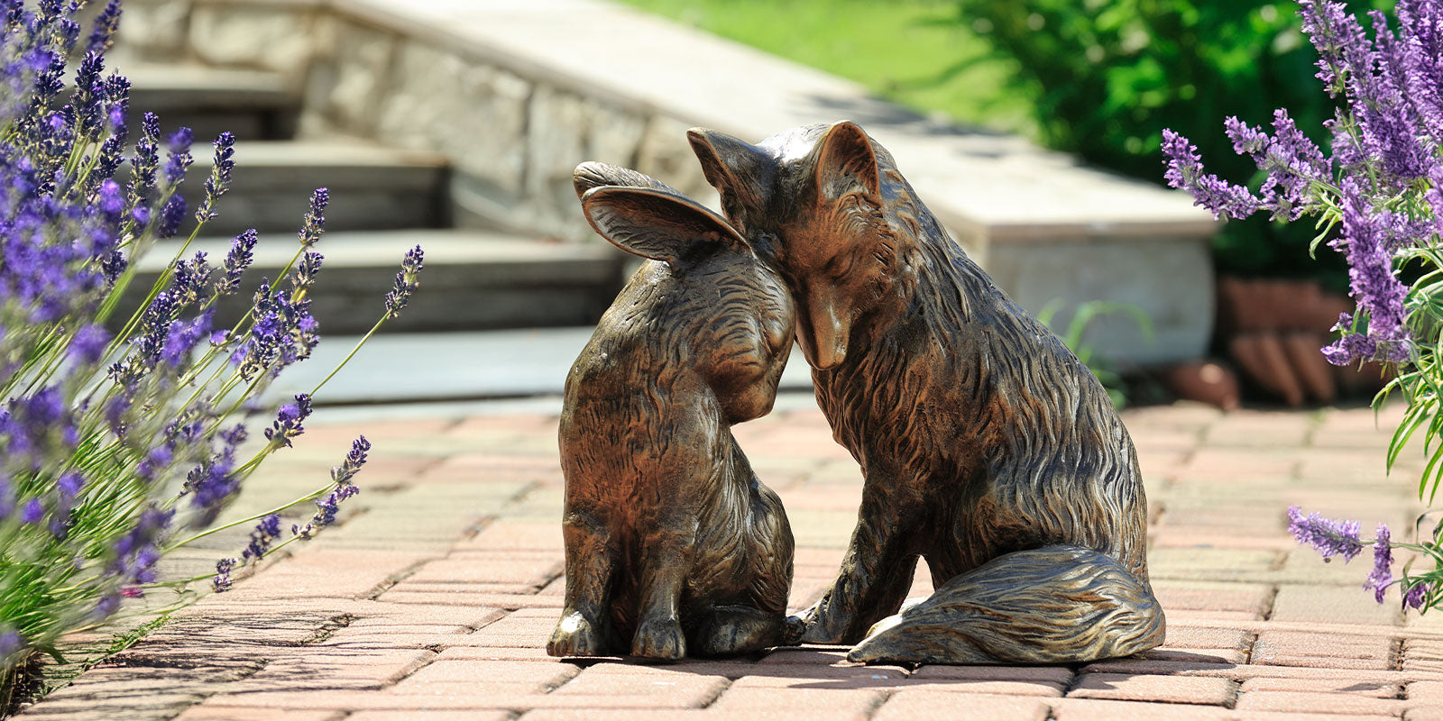 Curious Couple Garden Sculpture features a rabbit and fox nuzzling foreheads; they are shown on a brick path in a lavender garden