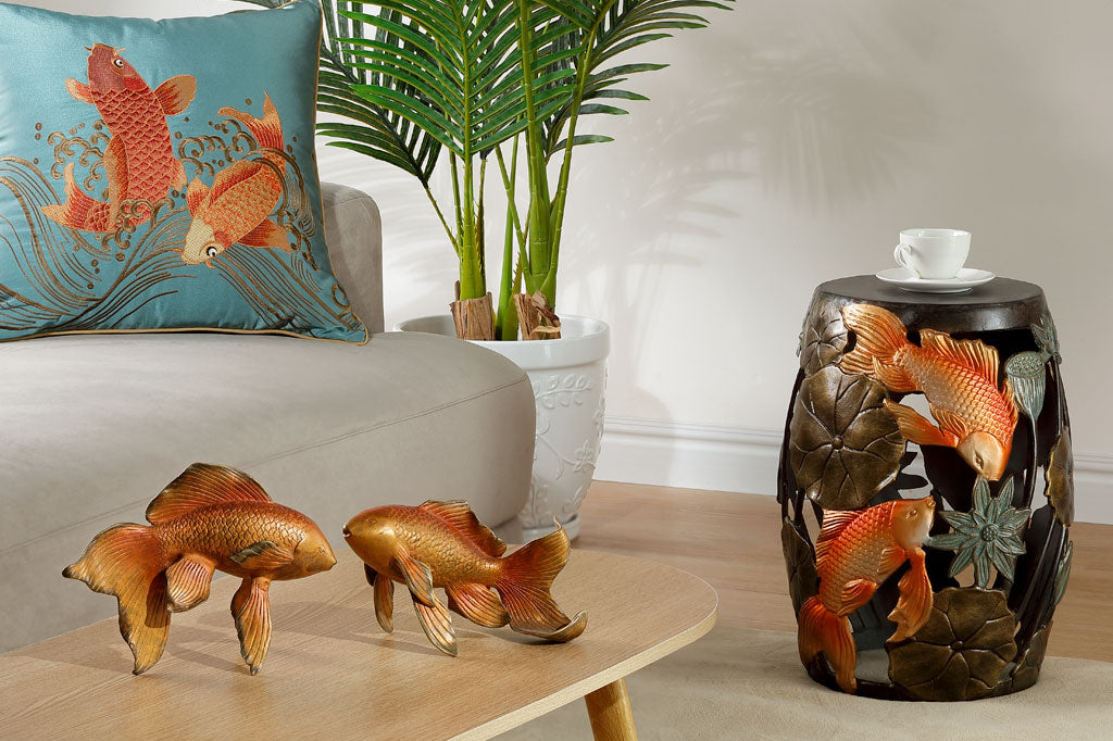 Cast Metal koi fish and water lily garden/ accent stool. golden orange koi surrounded by bronze water lilypad motifs. In a room with koi sculpture and koi satin pillow on a beige couch. Indoor palm houseplant in background.