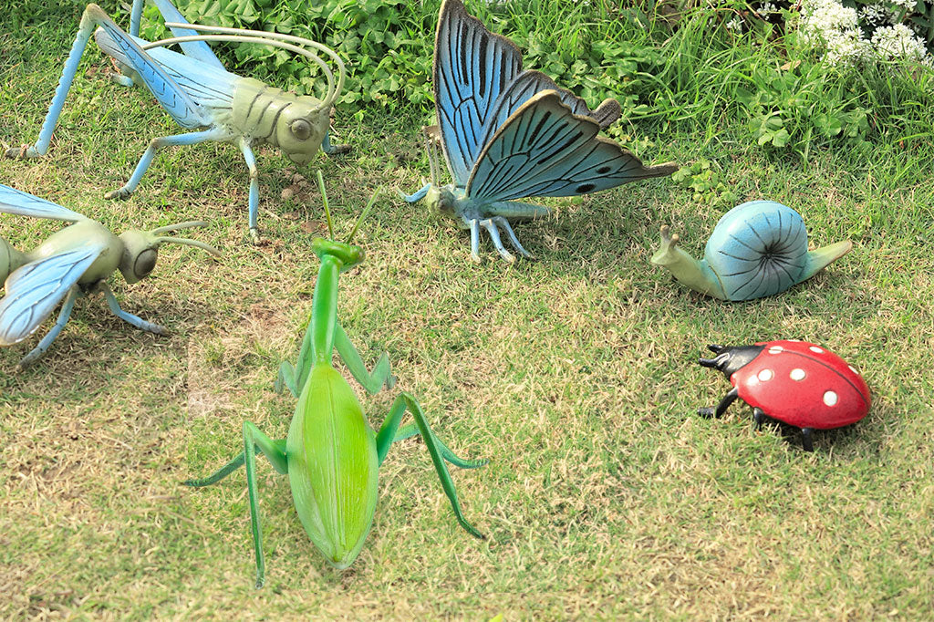 cast metal green garden sculpture of green praying manitis is shown with a group of insect sculptures on the lawn by a flower bed