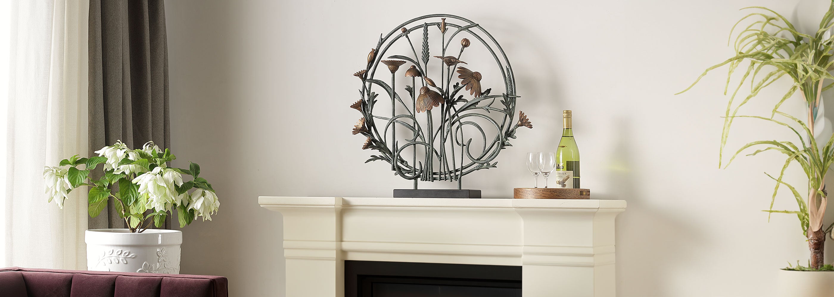 circular sculpture with rectancle base features cast metal arrangement of wildflowers, cone flowers and seed stalks; shown on fireplace mantle in living room