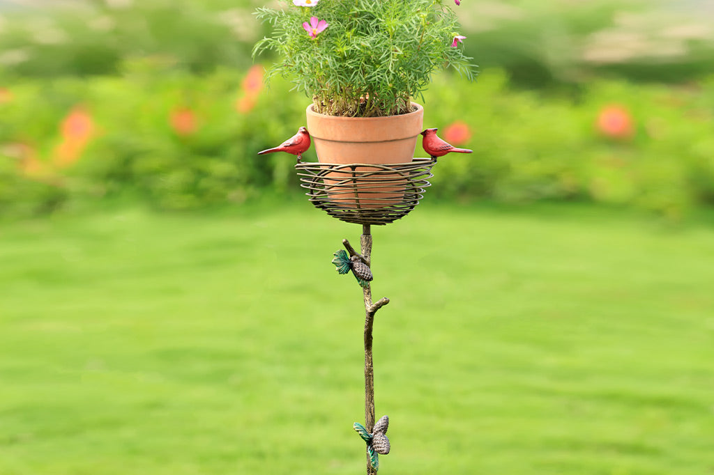 staked planter with red cardinals and a pine branch and pinecone motif adorns a bird nest used as a planter, shown in a garden setting