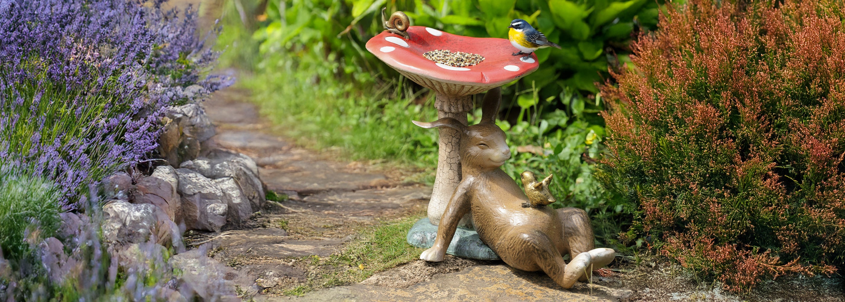 Garden birdbath sculpture shown in garden. shaped like bunny under a toadstool, on a stone path by lavender bushes