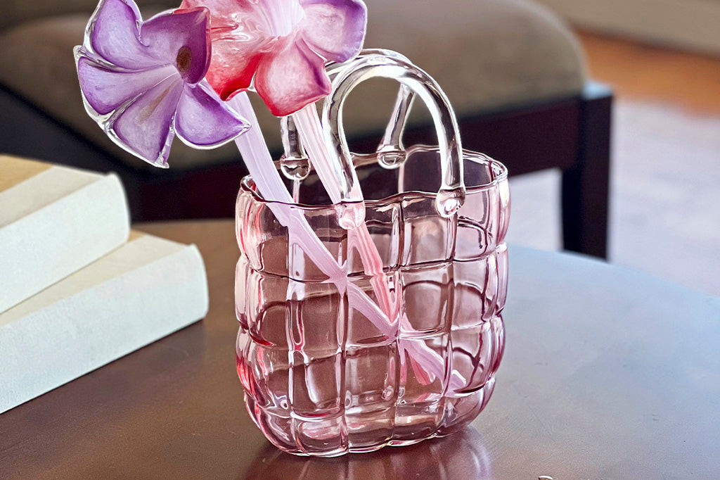 Pink glass vase shaped like handbag shown holding glass flower stems, displayed on coffeetable in living room with books and couch