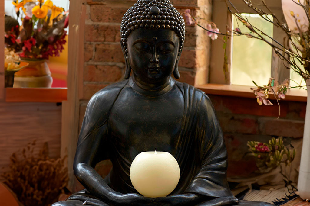 round LED candle shown in palms of buddha sculpture in rustic room with dried flowers around
