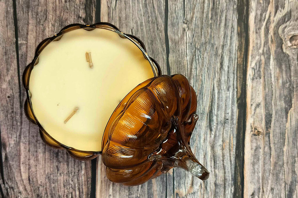 re-usable glass pumpkin candle with scent of pumpkin, vanilla, and nectarine shown on wood table; top view shows 3 wicks and the lid off to the side