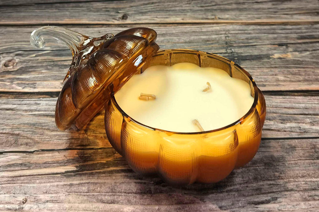 re-usable glass pumpkin candle with scent of pumpkin, vanilla, and nectarine shown on wood table. Lid is tilted to reveal the candle with 3 wicks