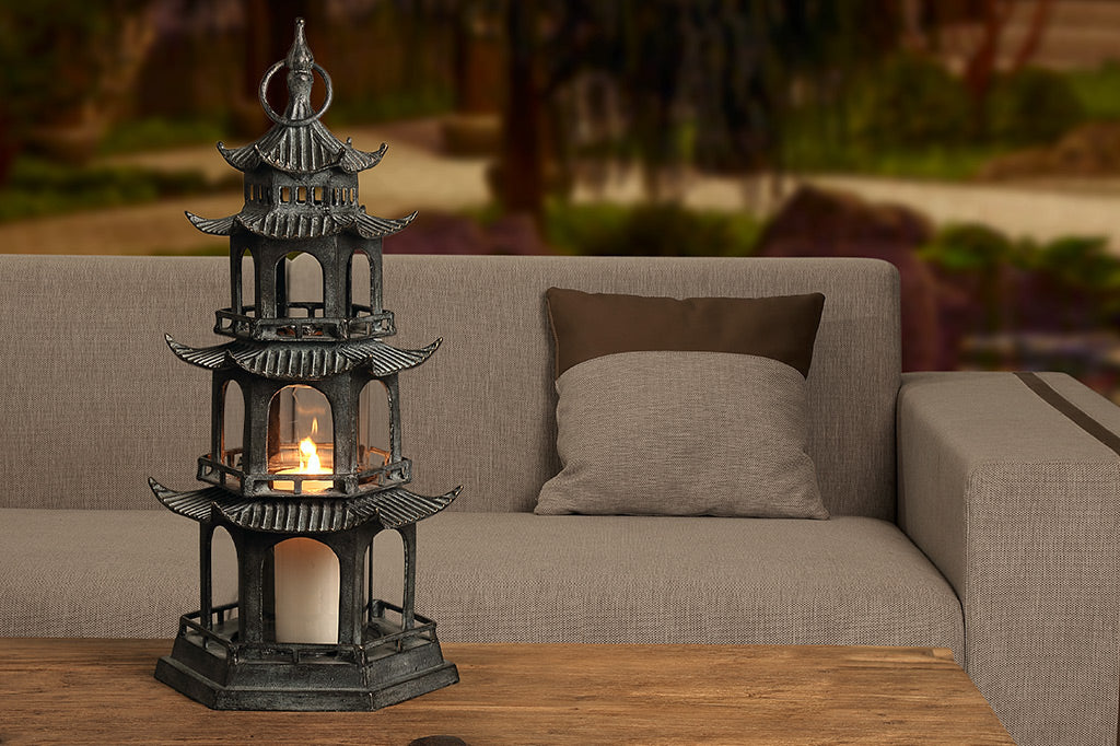 sculptural cast metal lantern shaped like Chinese pagoda holding pillar candle shown outside by sofa and trees at dusk