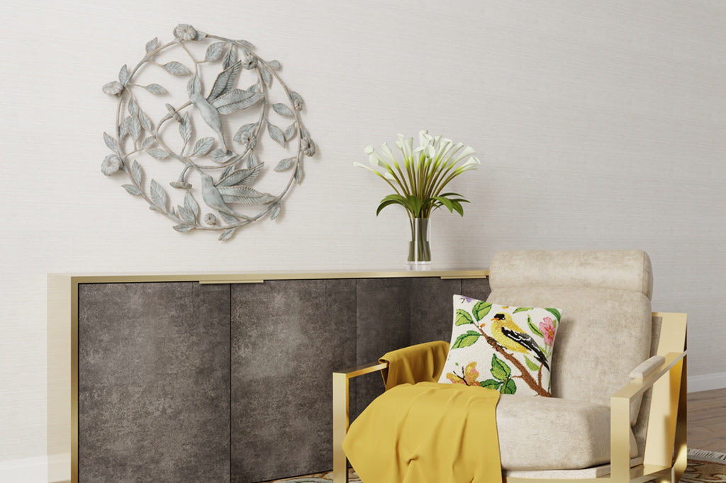Large circular wall art made from cast metal in a motif of flowering vines and 2 sipping hummingbirds. Wall art that hangs in a room over a console table, near a chair with throwblanket and pillow