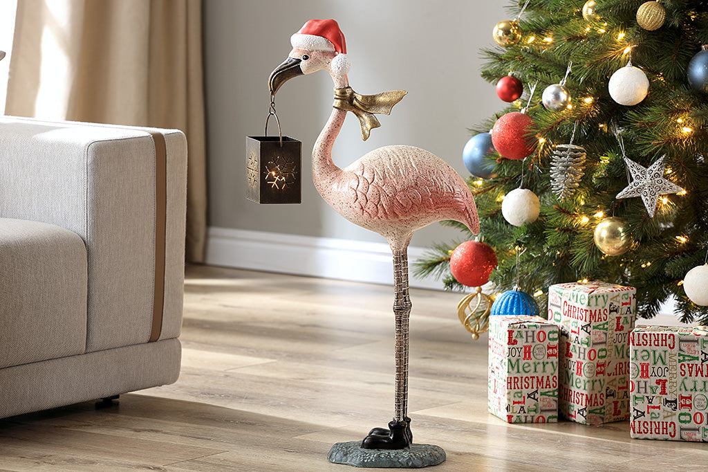 Flamingo holiday sculpture lantern shown with gold scarf and santa hat in red and white. Stagd in living room with christmas tree and couch