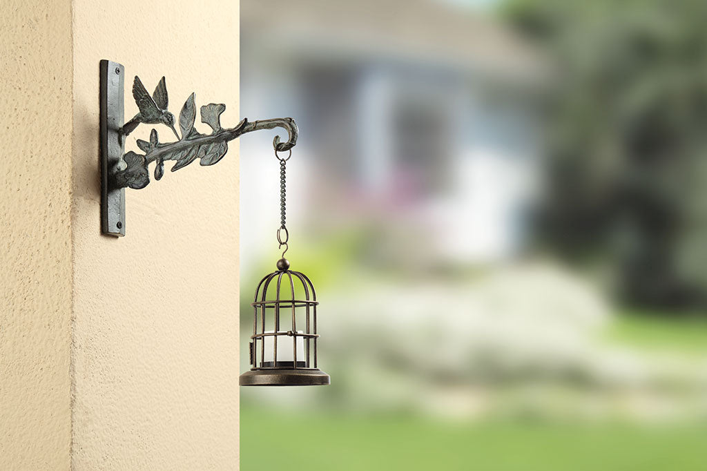 Hummingbird Mounted Hanger shown mounted outside of a house in a garden setting and holding a hanging votive lantern
