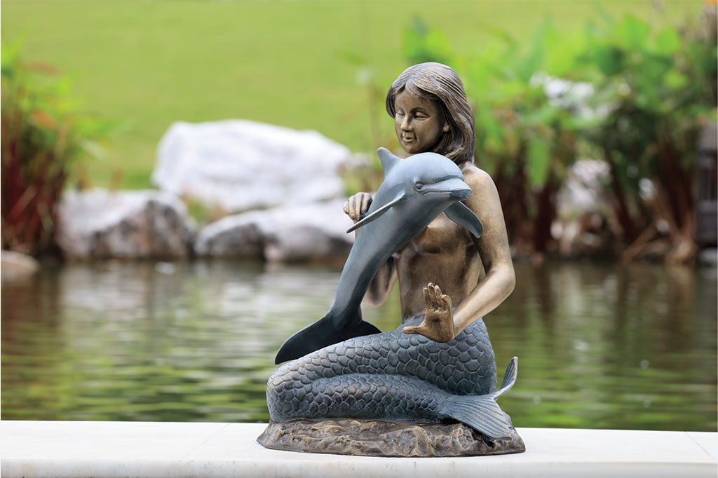 Bronze and verdi cast metal sculpture of a mermaid encountering a dolphin by the edge of a pond