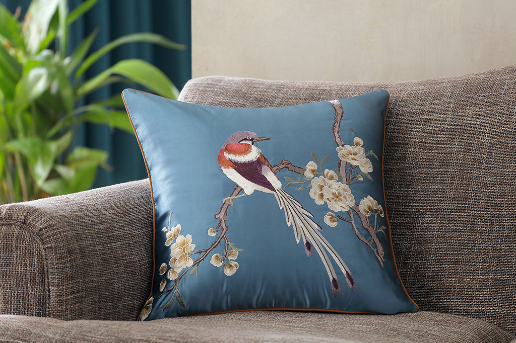 dark blue satin pillow with embroidered imagery of a bird perched on flowering branch placed on couch 
