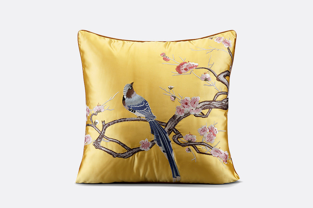 yellow satin pillow with embroidered imagery of blue flycatcher bird perched on blossoming branch against gray background 