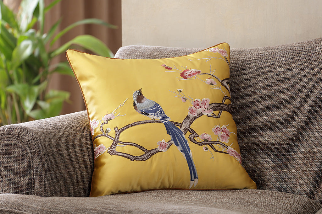 yellow satin pillow with embroidered imagery of blue flycatcher bird perched on blossoming branch propped up on couch 
