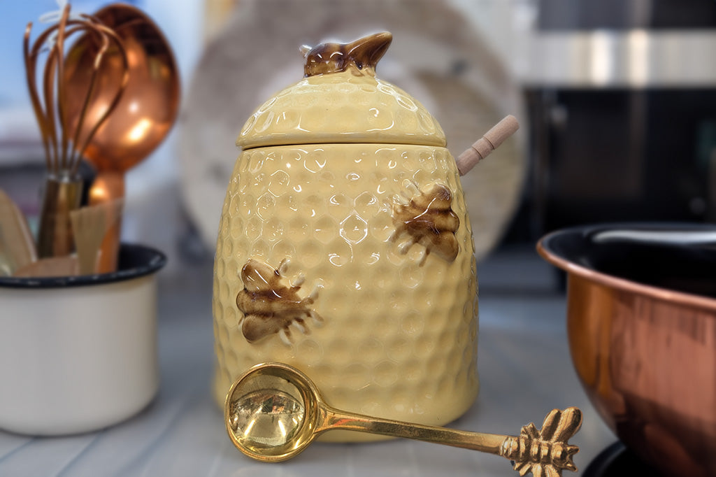 stoneware honeypot shaped like a beehive with wooden spoon, sitting in a kitchen with copper bowl, brass bee spoon, and assorted baking supplies