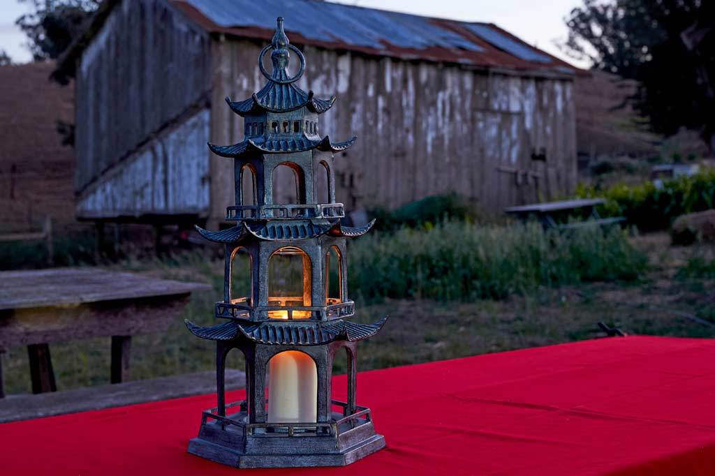 sculptural cast metal lantern shaped like Chinese pagoda holding pillar candle shown outside on a picnic table at dusk. A barn is in the background and the candlelight glows through the lantern temple windows.