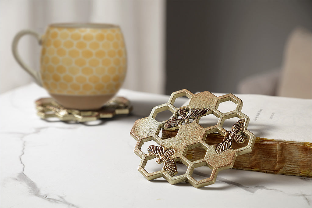 Single coaster shaped like honeycomb with bee accents shown propped up on book, mug in the background on matching coaster