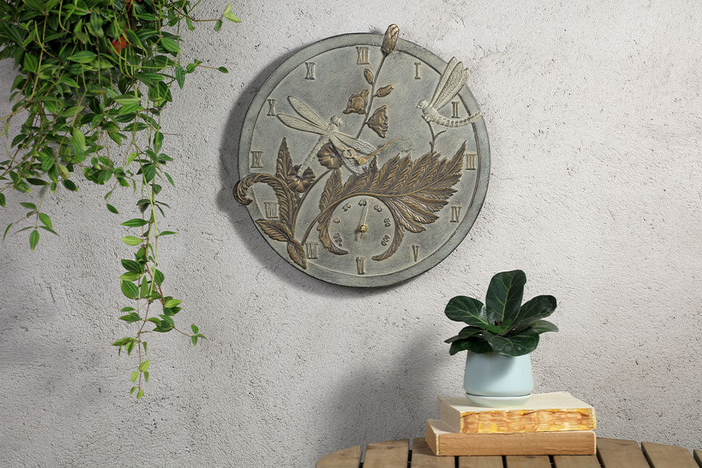 Metal sculpted wall clock and thermometer features fern flower, and dragonflies in zinc and bronze finishes. It is on a wall in a garden patio setting