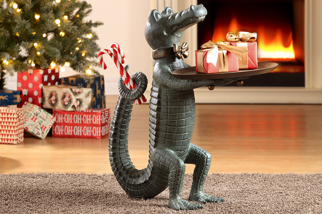 Holiday décor: Standing Crocodile dressed as a butler holds a serving tray whilst hiding candy canes behind its back. Shown on rug in front of fireplace near a Christmas tree and wrapped presents.